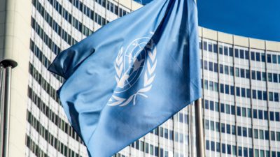 En blå flagga med FN:s symbol på. Flaggan hänger på en flaggstång framför en skyskrapa.